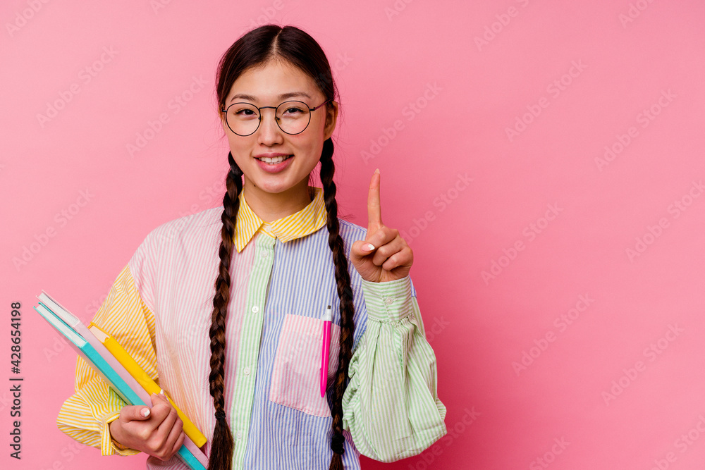 Young chinese student woman holding books wearing a fashion multicolour shirt and braid, isolated on pink background showing number one with finger. Young chinese student woman holding books wearing a fashion multicolour shirt and braid, isolated on pink background showing number one with finger.