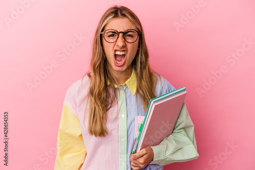 Young caucasian blonde student woman holding books isolated screaming very angry and aggressive.