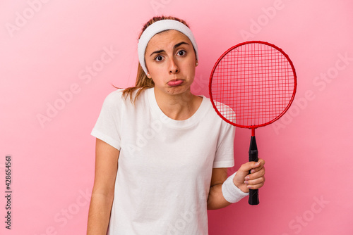 Young cute blonde caucasian woman holding a badminton racket isolated on pink background shrugs shoulders and open eyes confused.