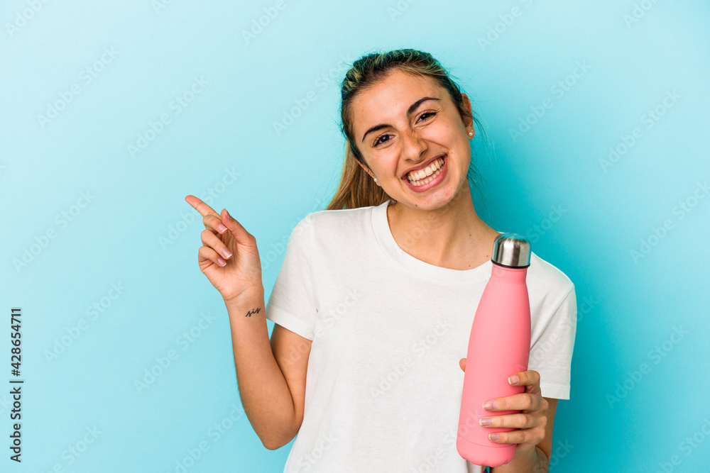Young blonde caucasian woman holding a thermo isolated on blue background smiling and pointing aside, showing something at blank space. Young blonde caucasian woman holding a thermo isolated on blue background smiling and pointing aside, showing something at blank space.