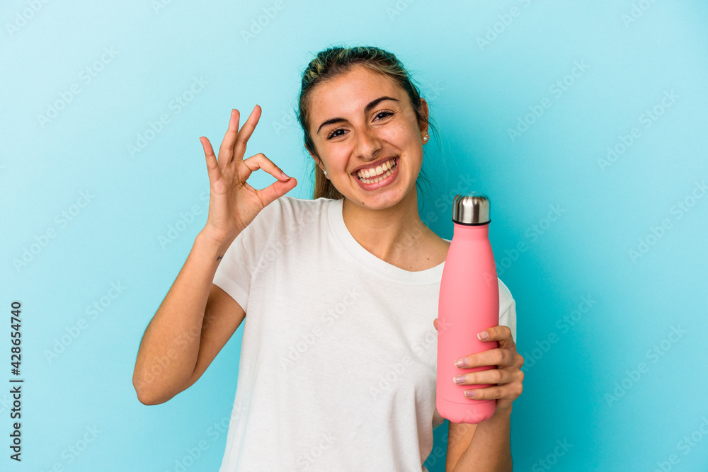 Young blonde caucasian woman holding a thermo isolated on blue background cheerful and confident showing ok gesture. Young blonde caucasian woman holding a thermo isolated on blue background cheerful and confident showing ok gesture.