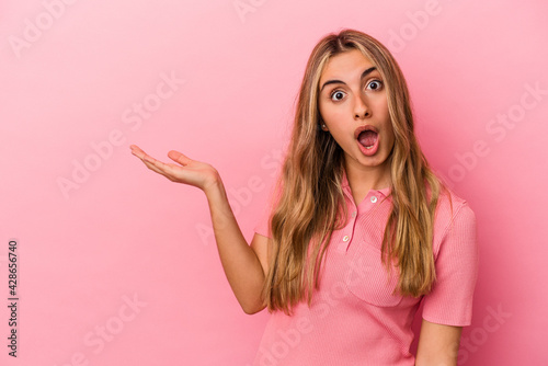 Young blonde caucasian woman isolated on pink background impressed holding copy space on palm.