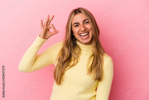 Young blonde caucasian woman isolated on pink background winks an eye and holds an okay gesture with hand.