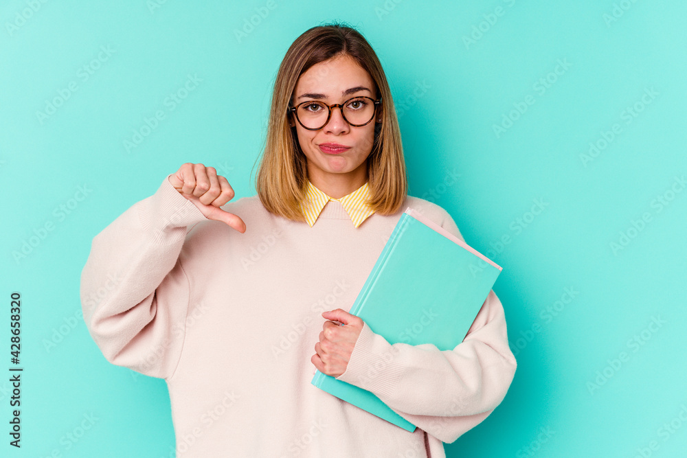 Young student woman holding books isolated on blue background showing a dislike gesture, thumbs down. Disagreement concept.
