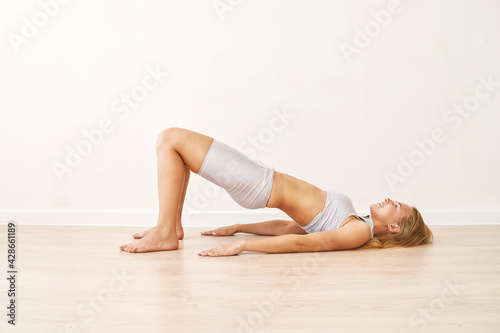 Young pretty woman standing in pelvic brigde. Yoga exercise at gym. Home sport workout. Elbows static balance stand. Floor stretching. Healthy lifestyle. Long blond hair. Smiling female person. Side photo