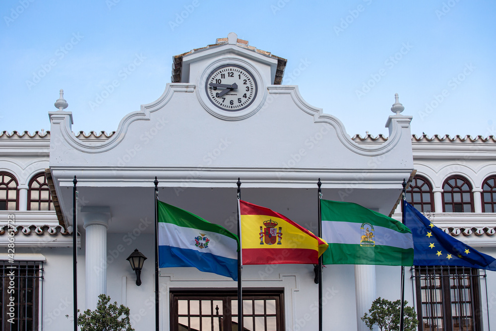 The flag of Spain, the flag of Andalusia, the flag of the European Union against the background of the faiths with a clock. Andalusia Mijas Pueblo - obrazy, fototapety, plakaty 