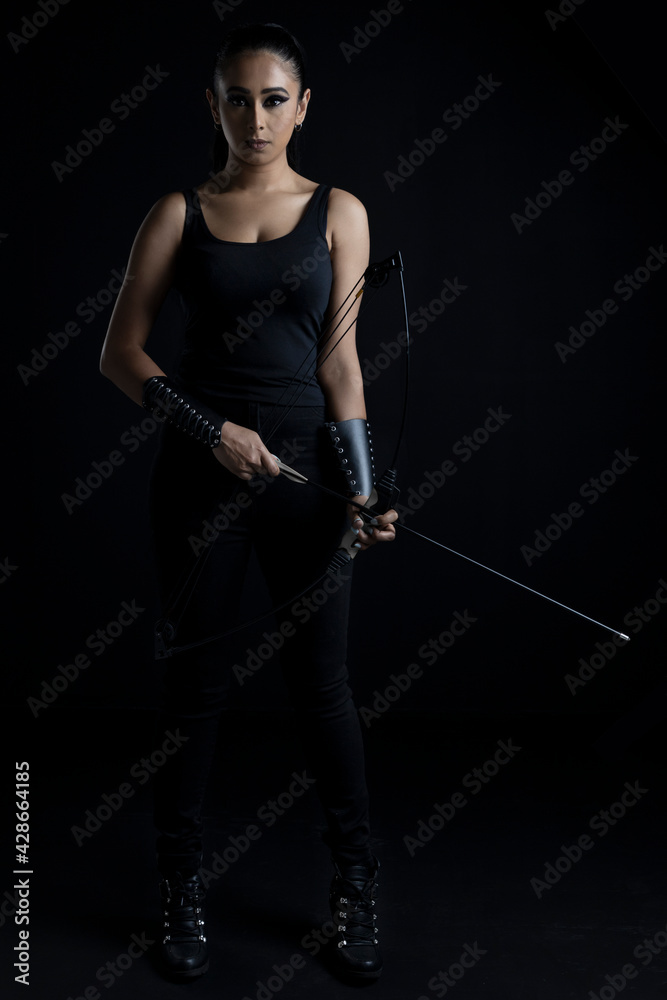 An attractive dark-haired woman in fantasy/urban fantasy style costume holding a bow and arrow and posing against a plain black backdrop