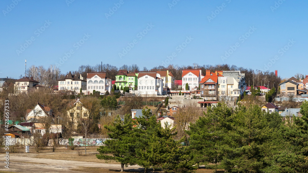 Village houses on a hill against a blue sky. Meadow with trees and grass in the field in front of the house. Horizontal village landscape with summer sunny day.