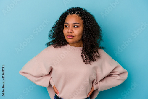 Young african american woman isolated on blue background looks aside smiling, cheerful and pleasant.