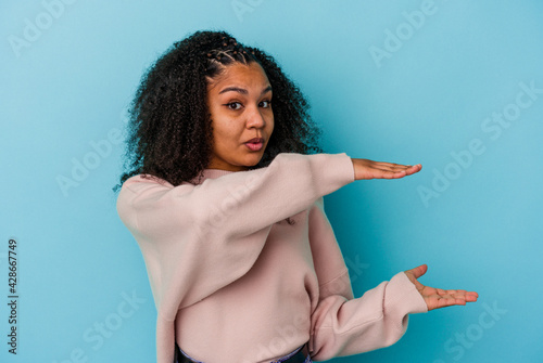 Young african american woman isolated on blue background holding something with both hands, product presentation.