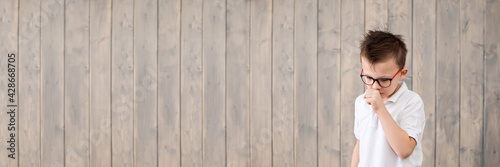 Cute little boy wearing glasses coughing, on brown wooden background. Copy space, extra wide banner. Place for text