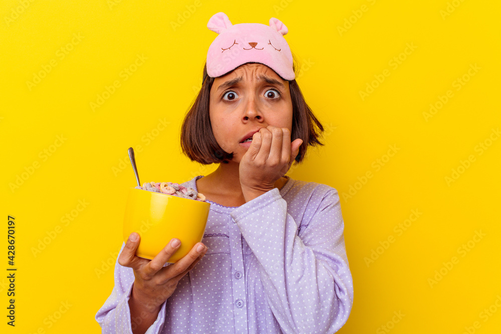 Young mixed race woman eating cereals wearing a pijama isolated on yellow background biting fingernails, nervous and very anxious. Young mixed race woman eating cereals wearing a pijama isolated on yellow background biting fingernails, nervous and very anxious.