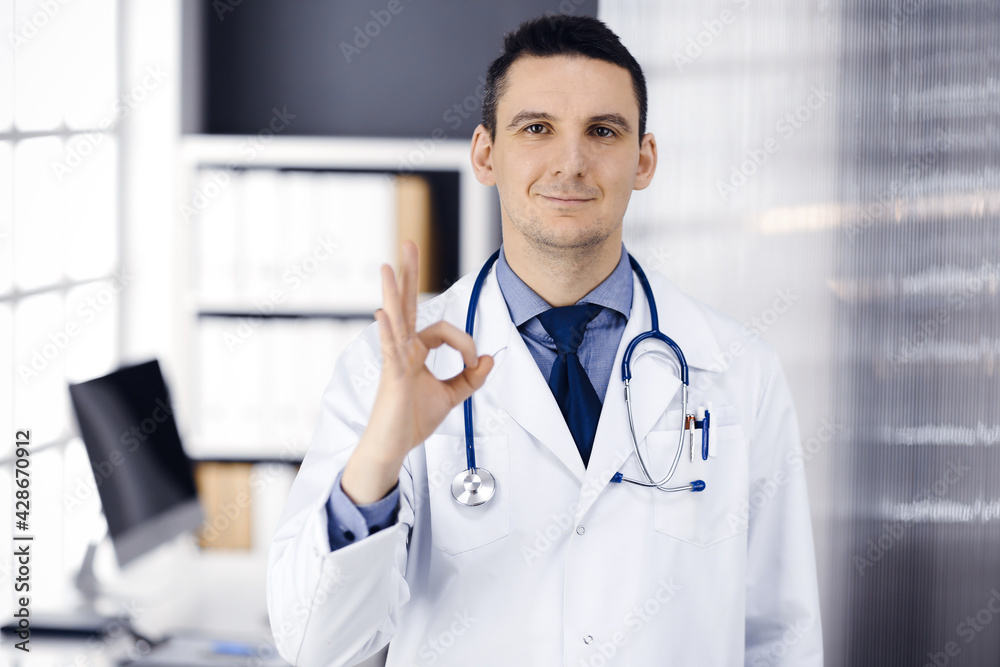 Friendly male doctor standing straight with OK sign in clinic near his working place, closeup. Perfect medical service in hospital. Medicine and healthcare concept Friendly male doctor standing straight with OK sign in clinic near his working place, closeup. Perfect medical service in hospital. Medicine and healthcare concept