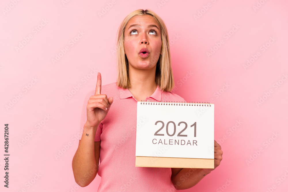 Young venezuelan woman holding a calendar isolated on pink background pointing upside with opened mouth. Young venezuelan woman holding a calendar isolated on pink background pointing upside with opened mouth.
