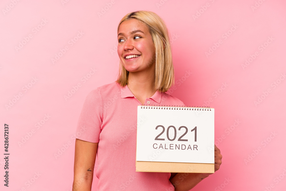 Young venezuelan woman holding a calendar isolated on pink background looks aside smiling, cheerful and pleasant. Young venezuelan woman holding a calendar isolated on pink background looks aside smiling, cheerful and pleasant.