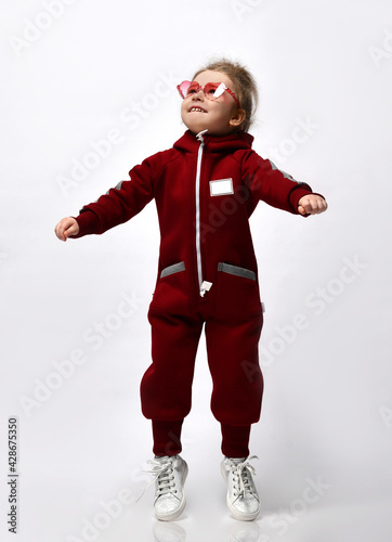 Cute little doshkolnitsa warm sportswear with a hood and white sneakers jumping with arms raised above the background with copy space. Studio portrait. Promotion of children's outerwear