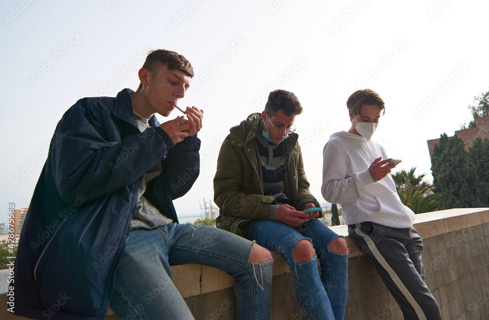 A closeup of three young Caucasian and Hispanic guys sitting outdoors smoking and using their phones wearing sanitary masks