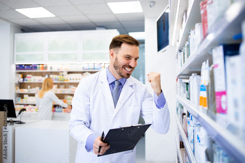 Positive caucasian pharmacist standing by the shelf with medicines in pharmacy store.