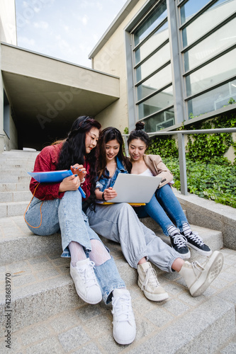 Group of Asian female students using a laptop and studying together while sitting on the stairs of the university campus.