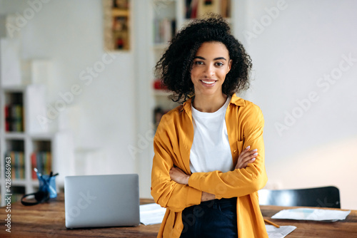 Portrait of a pretty African American girl. Confident stylish young african american woman with curly hair, stands near the desktop with crossed arms, looks at the camera, smiles friendly photo