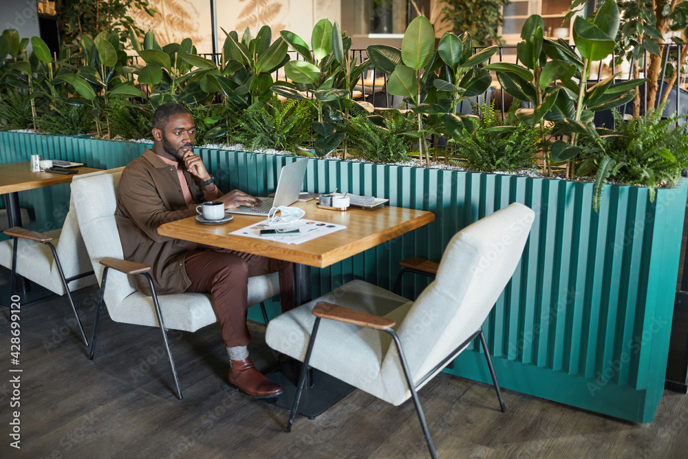 Full length portrait of successful African American businessman working with laptop while sitting at table in eco friendly green cafe interior, copy space Full length portrait of successful African American businessman working with laptop while sitting at table in eco friendly green cafe interior, copy space