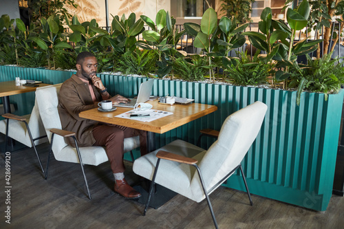 Full length portrait of successful African American businessman working with laptop while sitting at table in eco friendly green cafe interior, copy space