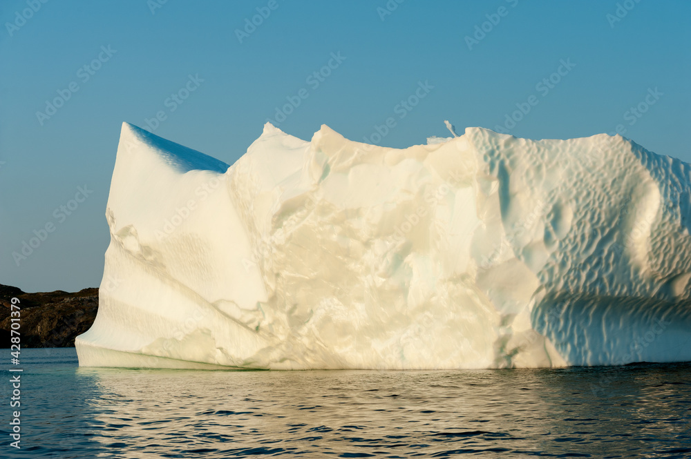 A large white iceberg formation floating in the cold ocean with layers of textured ice and snow. The ice is in transition melting from the warm rays of the sun at sunset. The berg is a blocky shape.