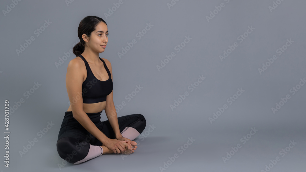 Joven mujer latina mexicana atleta instructora entrenando yoga dando calentamiento y estiramiento de musculos vestida de negro y haciendo ejercicio con fondo gris en pandemia por covid 19 coronavirus