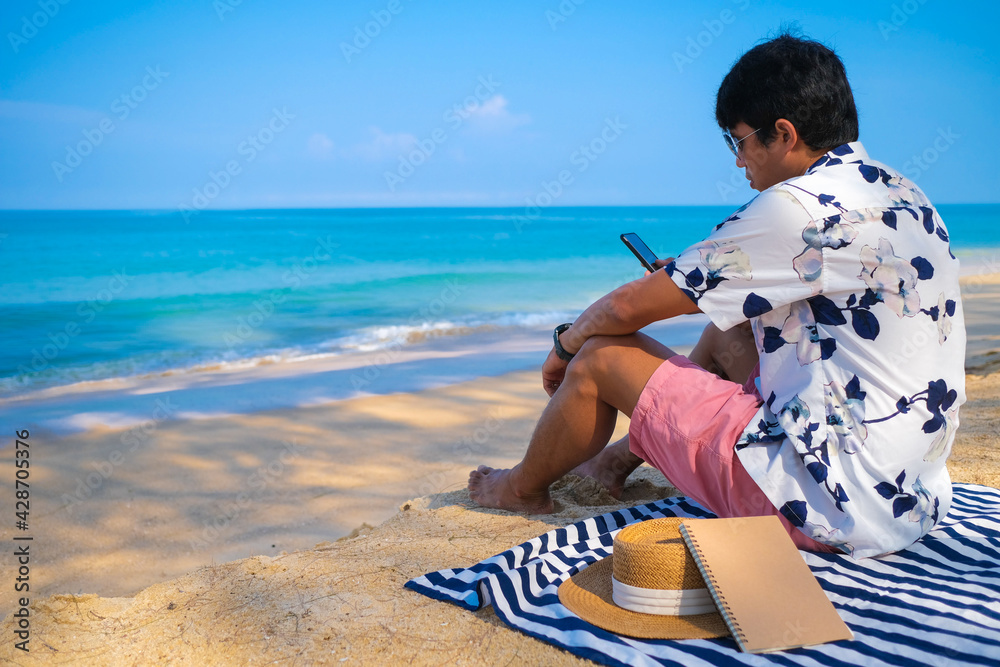 Asian man using smartphone and relaxing on the on striped beach blanket on the white sand beach. Summer vacation, summer in holiday concept. 