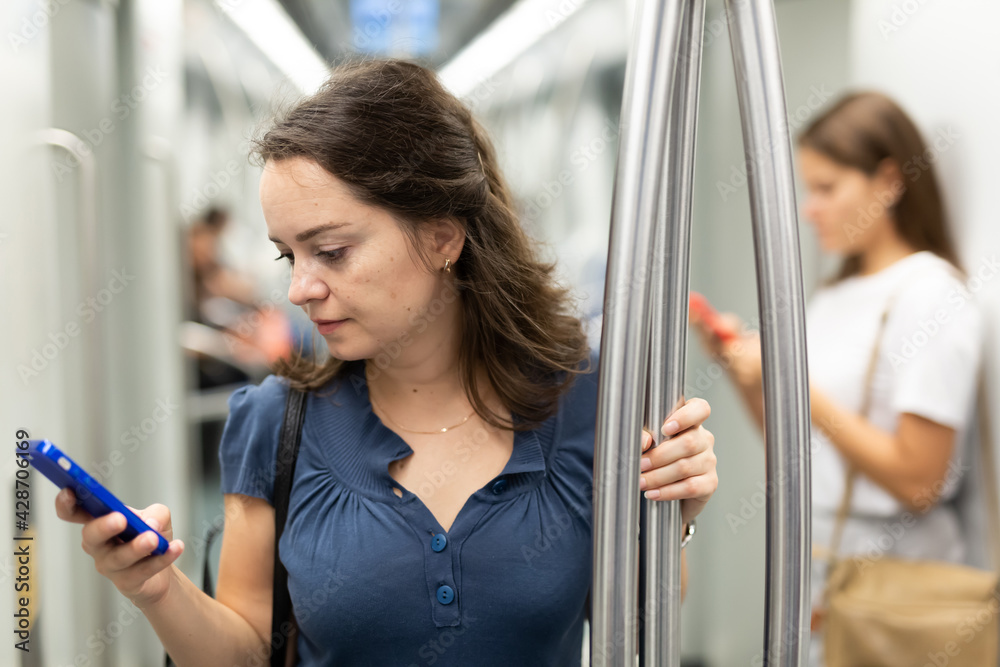 Young focused woman standing in subway car holding on handrails, browsing in her smartphone..