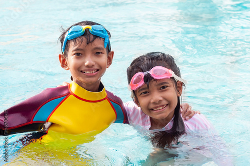 Asian brother and sister wearing a swimming suit and goggles looking and smiling with happy fun in the pool in the summer holiday. © Pannarai