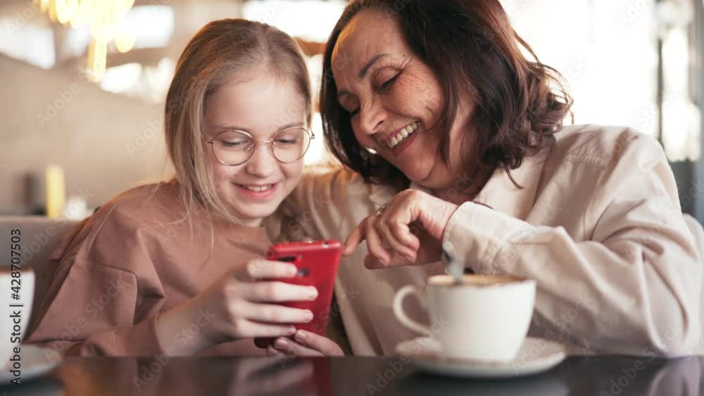 Happy grandmother and cute granddaughter using mobile phone together, while sitting at the cafe. Smiling senior grandmother and little girl having fun spending time together. Happy grandmother and cute granddaughter using mobile phone together, while sitting at the cafe. Smiling senior grandmother and little girl having fun spending time together.
