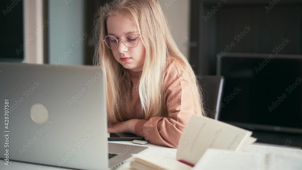 Beautiful 10 years old blond girl in glasses sitting at the desk doing her homework using a laptop. Beautiful 10 years old blond girl in glasses sitting at the desk doing her homework using a laptop.