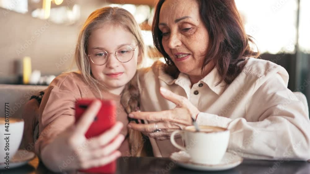 Happy grandmother and cute granddaughter taking selfies on the phone together, while sitting at the cafe. Cheerful grandmother with little child plays making photo on a mobile phone. Happy grandmother and cute granddaughter taking selfies on the phone together, while sitting at the cafe. Cheerful grandmother with little child plays making photo on a mobile phone.