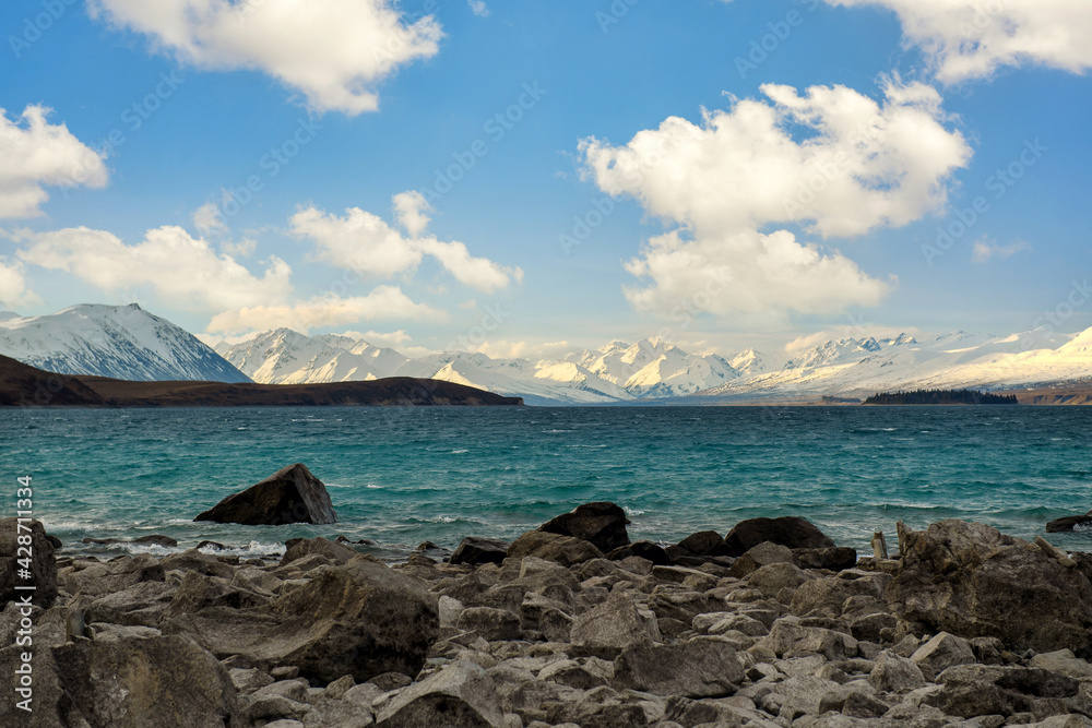 Rocks in turquoise water, waves in Lake Tekapo, background is Mount Cook Mountains. On a beautiful cloudy day on the South Island of New Zealand.