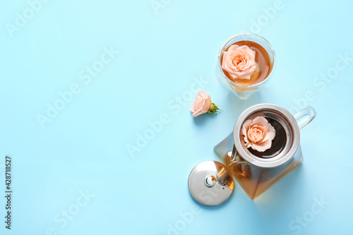 Teapot with hot tea, glass and rose flowers on color background