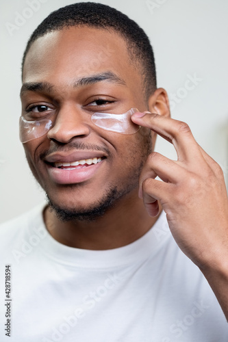 Man facial care. Skin rejuvenation. Cosmetic product advertising. Satisfied African guy in white t-shirt applying collagen gel face eye patches smiling on light background.