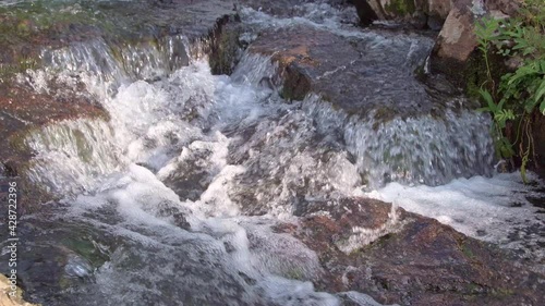 Forest stream cascades over granite boulders. Sun shines on bubbling water. Rocks and moss in riverbed. On shore, green grass moves with wind