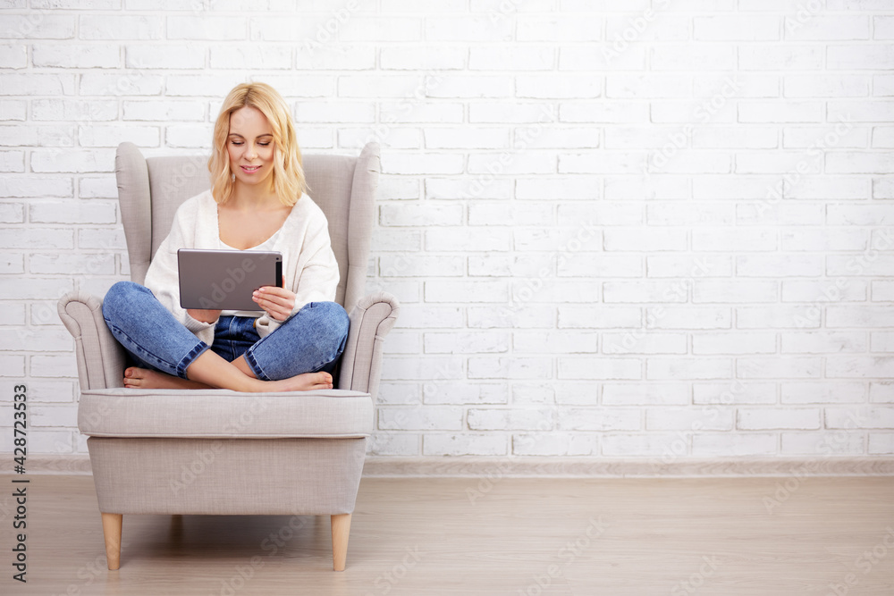 portrait of young beautiful woman sitting in armchair and using tablet pc - copy space over white brick wall portrait of young beautiful woman sitting in armchair and using tablet pc - copy space over white brick wall