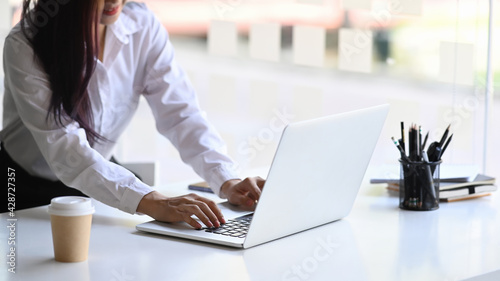 Cropped shot of smiling young female employee working with computer laptop at white office desk near window.