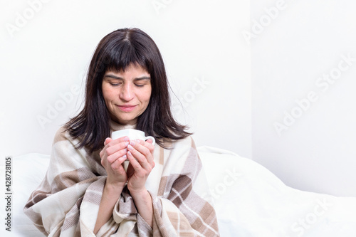Portrait of a sick attractive woman, wrapped in a blanket, holding a hot tea or coffee mug with steam