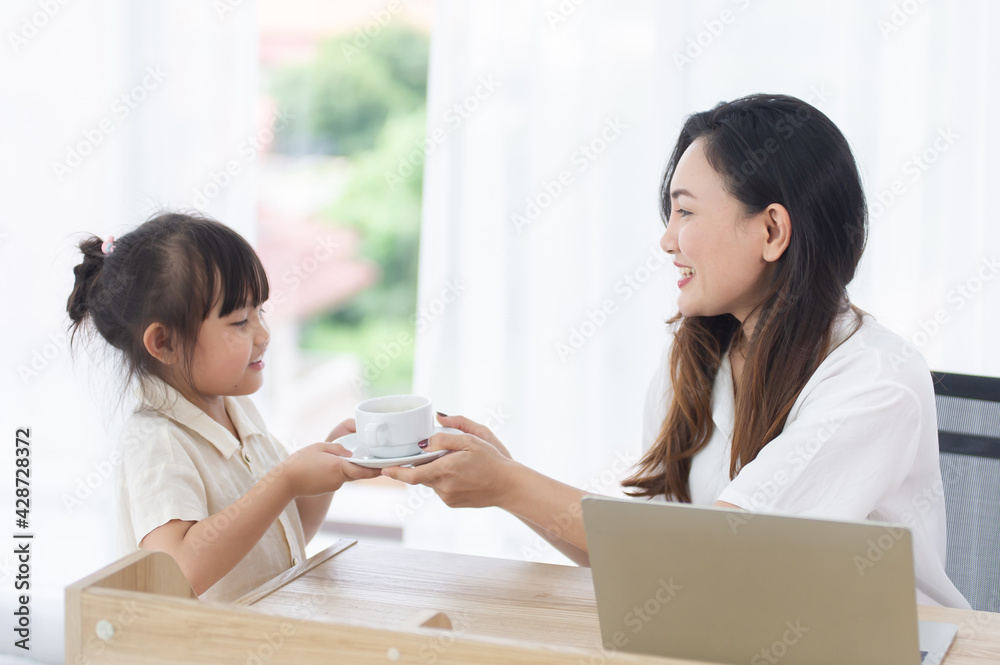 Cute happy little daughter give coffee to her smiling mother While mom works on computer laptop at home on holiday Cute happy little daughter give coffee to her smiling mother While mom works on computer laptop at home on holiday