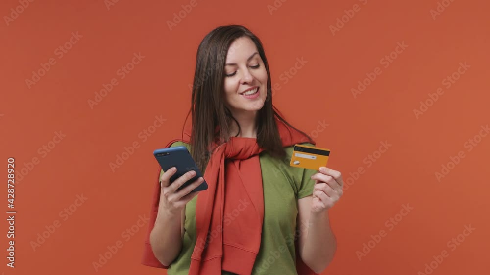 Young woman in basic casual olive green khaki t-shirt use mobile cell phone hold in hand credit bank card doing online shopping order delivery to home booking tour isolated on bright orange background Young woman in basic casual olive green khaki t-shirt use mobile cell phone hold in hand credit bank card doing online shopping order delivery to home booking tour isolated on bright orange background