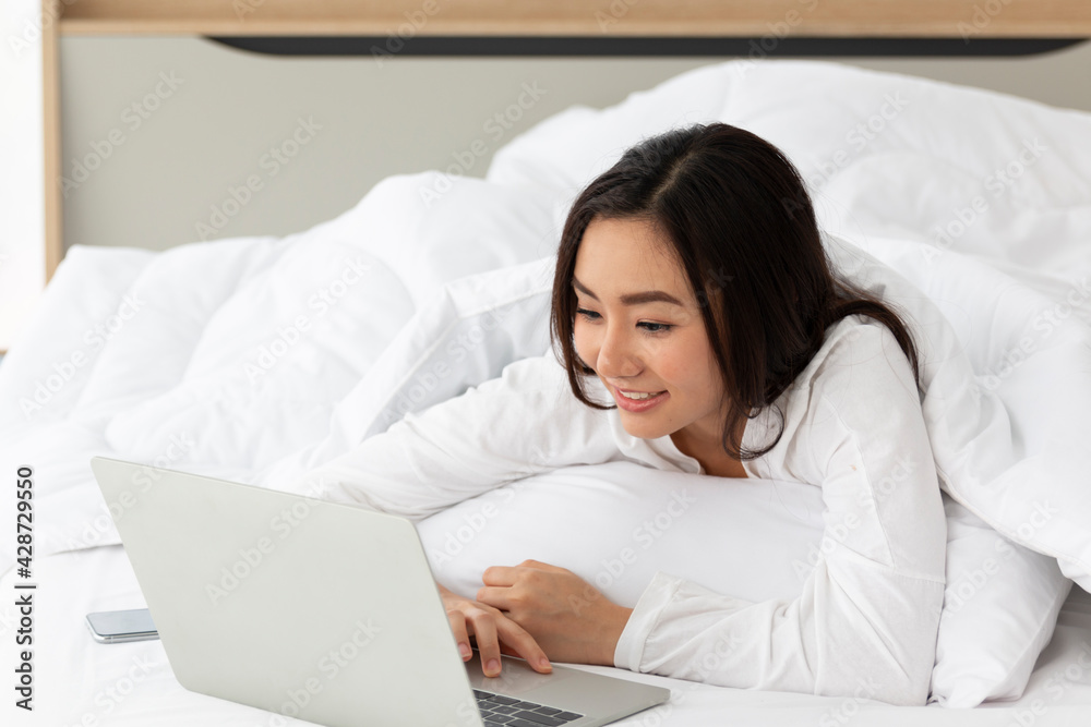 Young Asian female laying on bed using notebook computer. Happy female using laptop at home in bedroom. Working from home in quarantine lockdown. Women watching movie on laptop lying in bed. Young Asian female laying on bed using notebook computer. Happy female using laptop at home in bedroom. Working from home in quarantine lockdown. Women watching movie on laptop lying in bed.