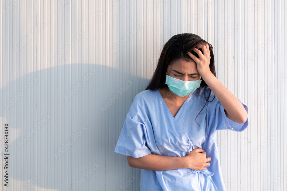 Female patient wearing mask standing by the wall feeling pain and depression. Young women recovering from surgery. Young Asian patient women in hospital, depressed and sad look by the wall. Female patient wearing mask standing by the wall feeling pain and depression. Young women recovering from surgery. Young Asian patient women in hospital, depressed and sad look by the wall.