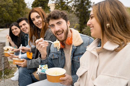 Young people with face mask on the wrist having fun eating take away food at the park using compostable and plastic free bowls and cutlery. New normal concept with people having fun together outdoor.