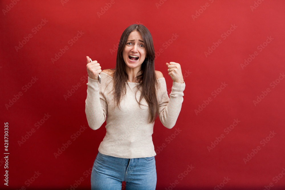 Angry Young beautiful caucasian girl wearing beige jacket and jeans over red background holding phone, Angry Young beautiful caucasian girl wearing beige jacket and jeans over red background holding phone,