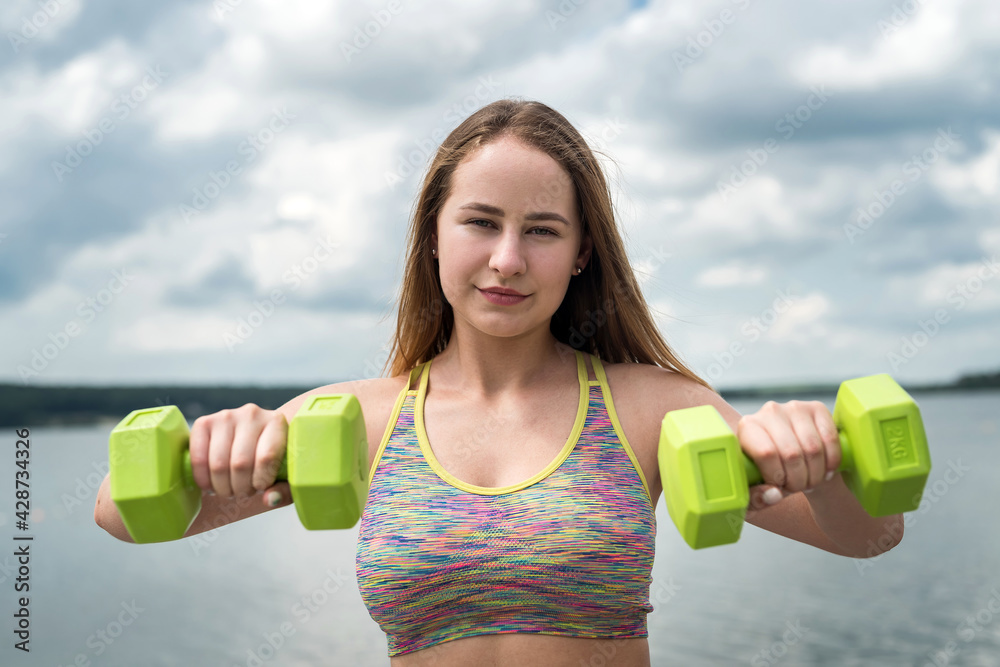 Young  fitness athletic trener doing exercises with dumbbells on beach lake. - obrazy, fototapety, plakaty 