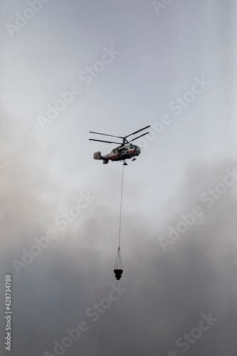 Firefighter helicopter with a water tank in smoke from a fire. Swirling smoke from a large fire.