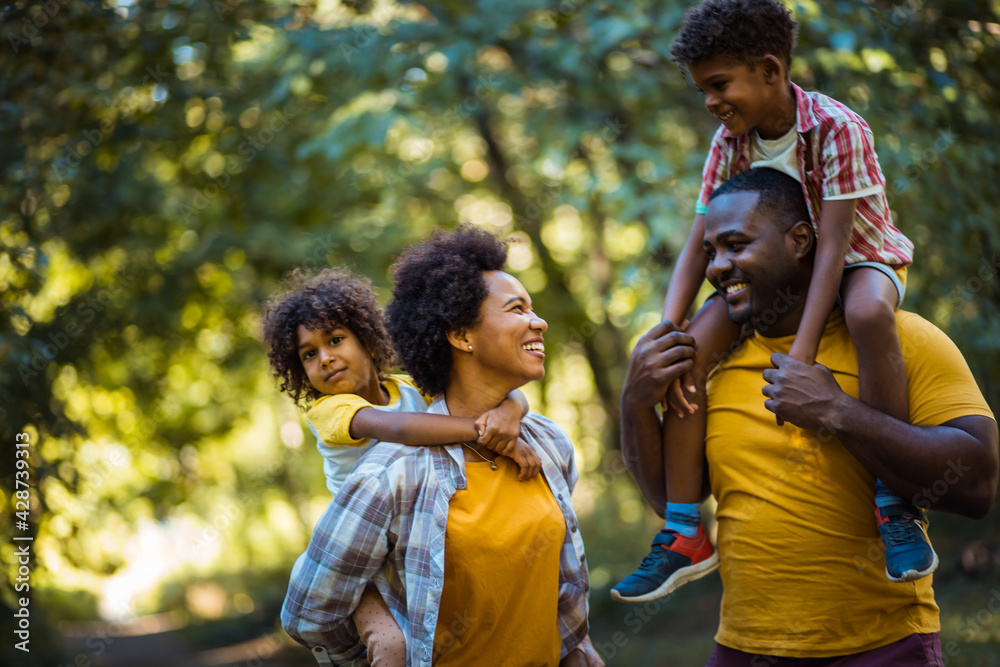 African American family walking trough park. Parents carrying children on piggyback. African American family walking trough park. Parents carrying children on piggyback.